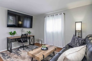 Una sala de estar moderna con una mesa de centro de madera, un sofá de cuero negro, un escritorio con una silla y un ventanal con cortinas.
