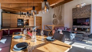 Una cocina moderna con armarios de madera, un comedor con mesa de madera dispuesta para una comida, y una sala de estar con televisor y asientos.