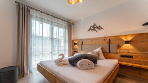 Un dormitorio moderno con cabecera de madera, un ventanal con cortinas de gasa, una cama con toallas ordenadas y una lámpara de pared.