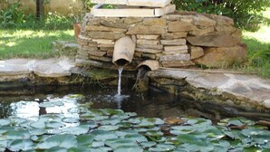 Ein Teich mit Seerosen und einer rosa Wasserlilie, eine Steinarchitektur mit einem Rohr sowie eine Steinsicht im Hintergrund.