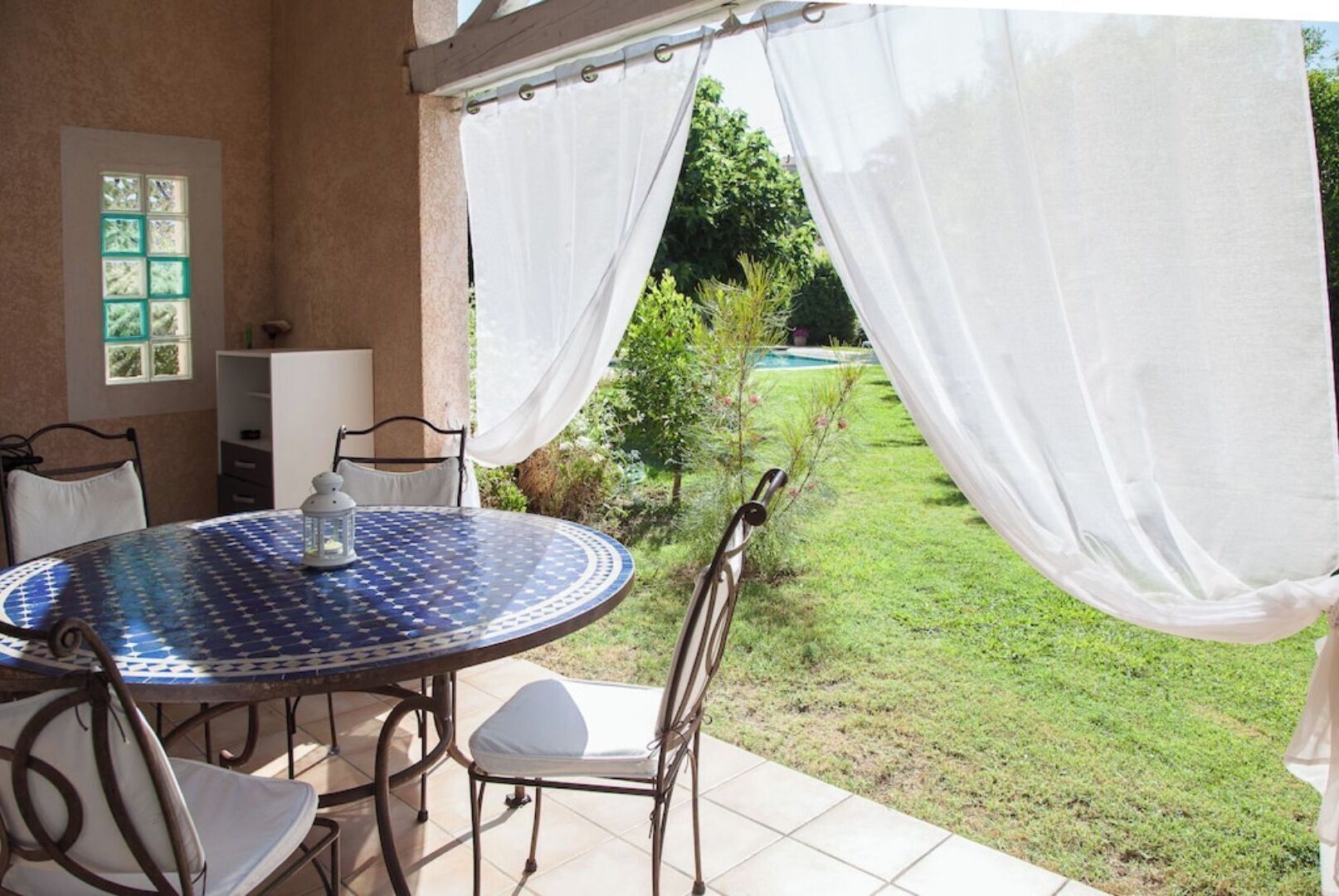 A patio with a round table and chairs, a white curtain, and a view of a grassy area with trees and a pool.