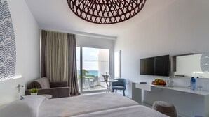 A modern hotel room with a large bed, a desk with a TV, a small table with a chair, and a balcony with a view. - MARGADINA HOTEL (Ayia Napa)