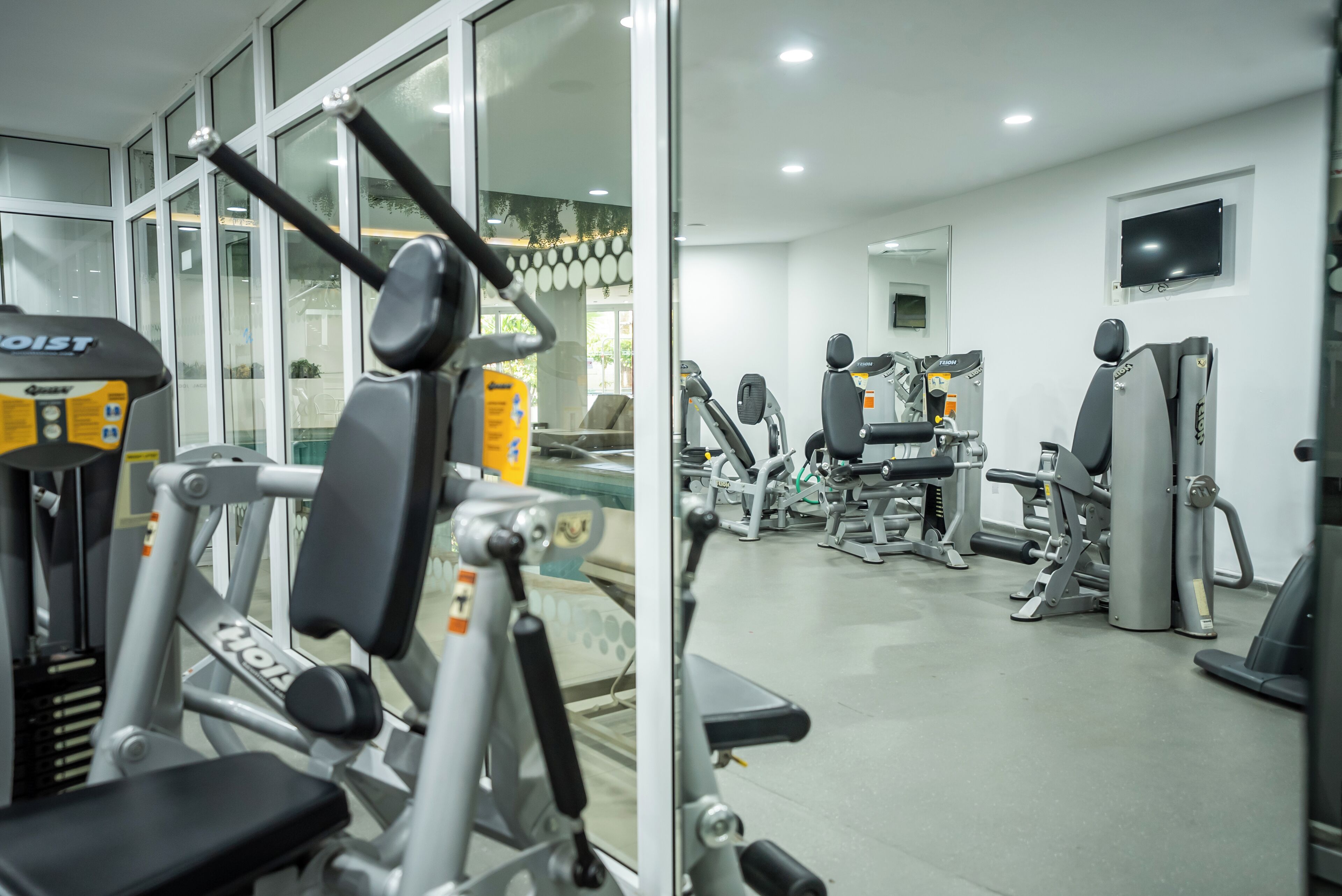 A gym with various exercise machines, a television mounted on the wall, and large windows allowing natural light.