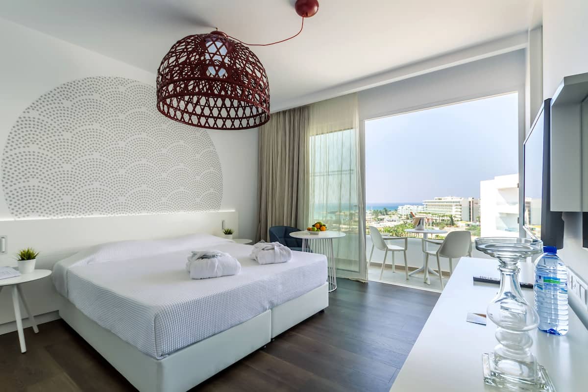 A modern hotel room with a large bed, a desk, a chair, a water bottle, and a view of the city.