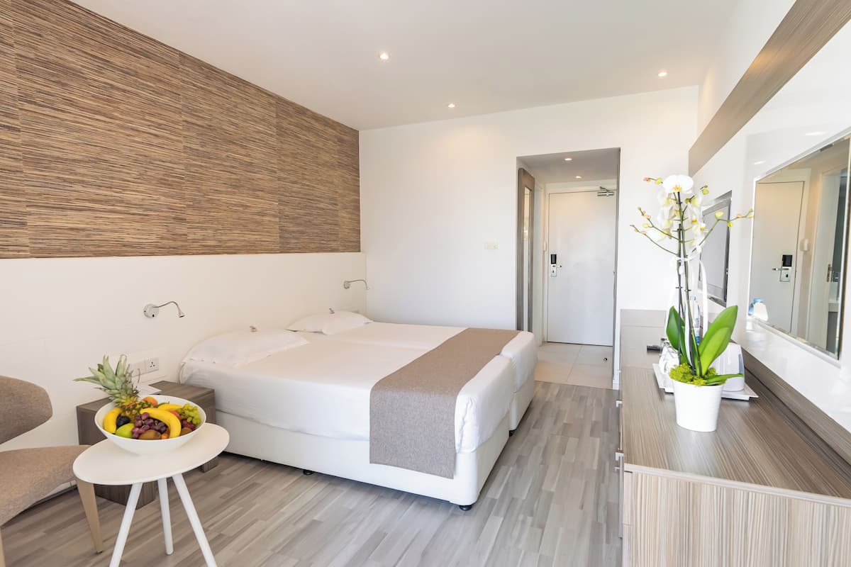 A modern hotel room with a large bed, a small bedside table with a fruit bowl, a potted plant, and a mirror above the bed.