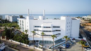 A modern building with a white facade, multiple floors, and a prominent 'margaritina' sign. It features a parking lot with several cars and palm trees around. - MARGADINA HOTEL (Ayia Napa)