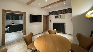 Un coin repas avec une table ronde en bois et quatre chaises marron, un téléviseur fixé au mur et une cuisine visible à travers une porte ouverte.