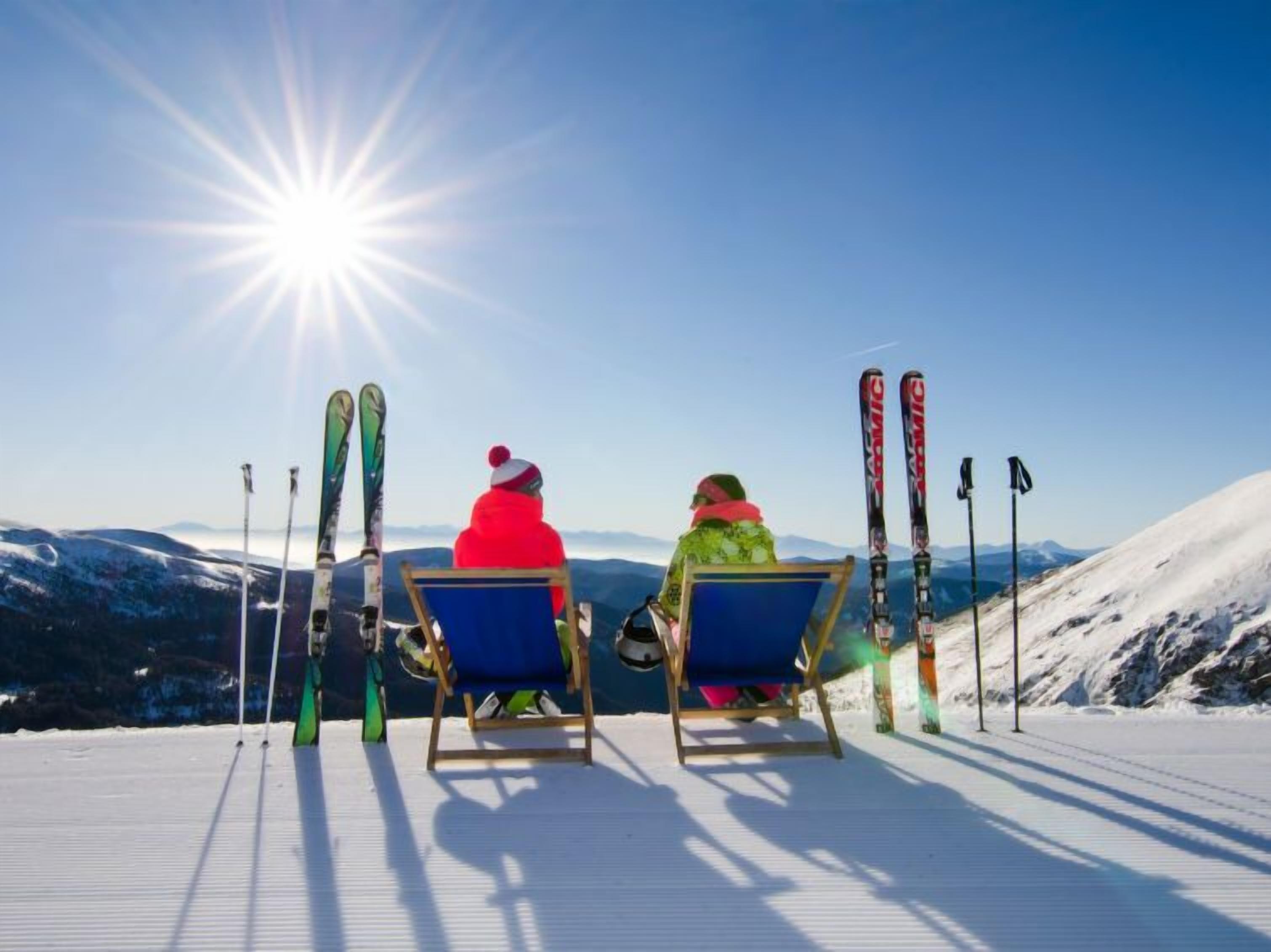 Two people sitting on chairs facing a snowy mountain slope with skis leaning against a pole.