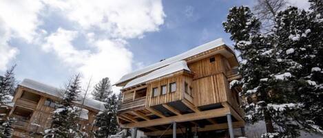 A wooden chalet with a sloped roof, elevated on stilts, surrounded by snow-covered trees.