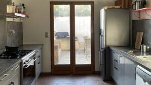 A modern kitchen with a gas stove, oven, and refrigerator. There is a wooden door leading to an outdoor area with a patio and chairs. - Mas Des Fleurs - Heated pool - Child friendly gardens - Beach 10mins   (Perpignan)