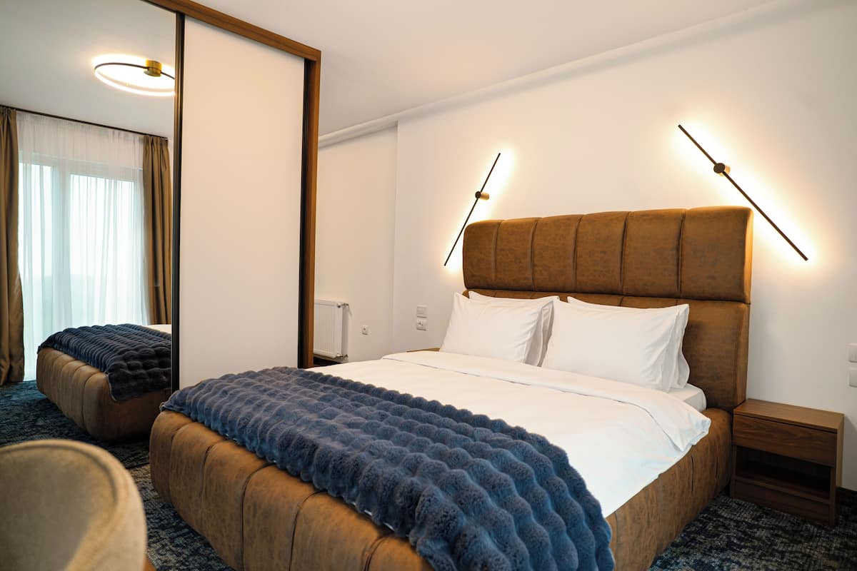 A hotel room with a large bed, a wooden headboard, a nightstand, a mirror, and a window with curtains.