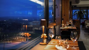A dining area with a table set for two, a view of the cityscape through large windows, and a potted plant on the windowsill. - Alexander Resort (Belgrade)