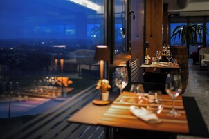 Un comedor con una mesa dispuesta para dos, vistas de la ciudad a través de amplias ventanas y una planta en maceta en el alféizar.