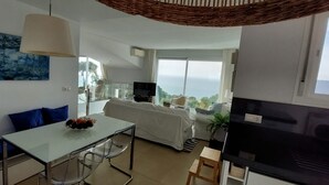 A modern living room with a glass dining table, a sofa, a TV, and a large window offering a view of the sea.