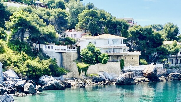 A coastal beach with clear turquoise water, rocky shoreline, and buildings in the background.