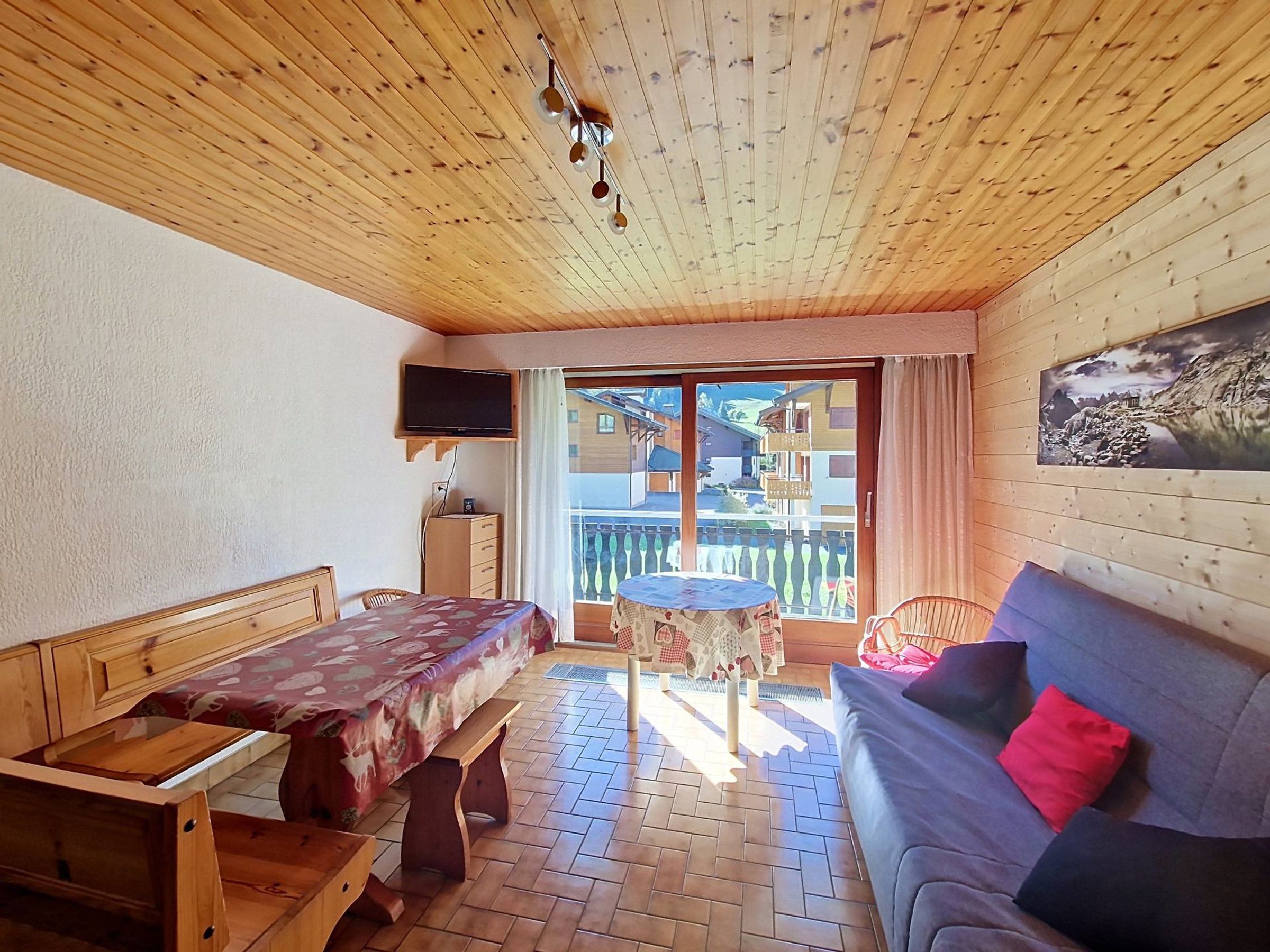 Una habitación con techo de madera, una cama con colcha estampada, un sofá con cojines, una pequeña mesa con mantel a cuadros y un televisor montado en la pared.