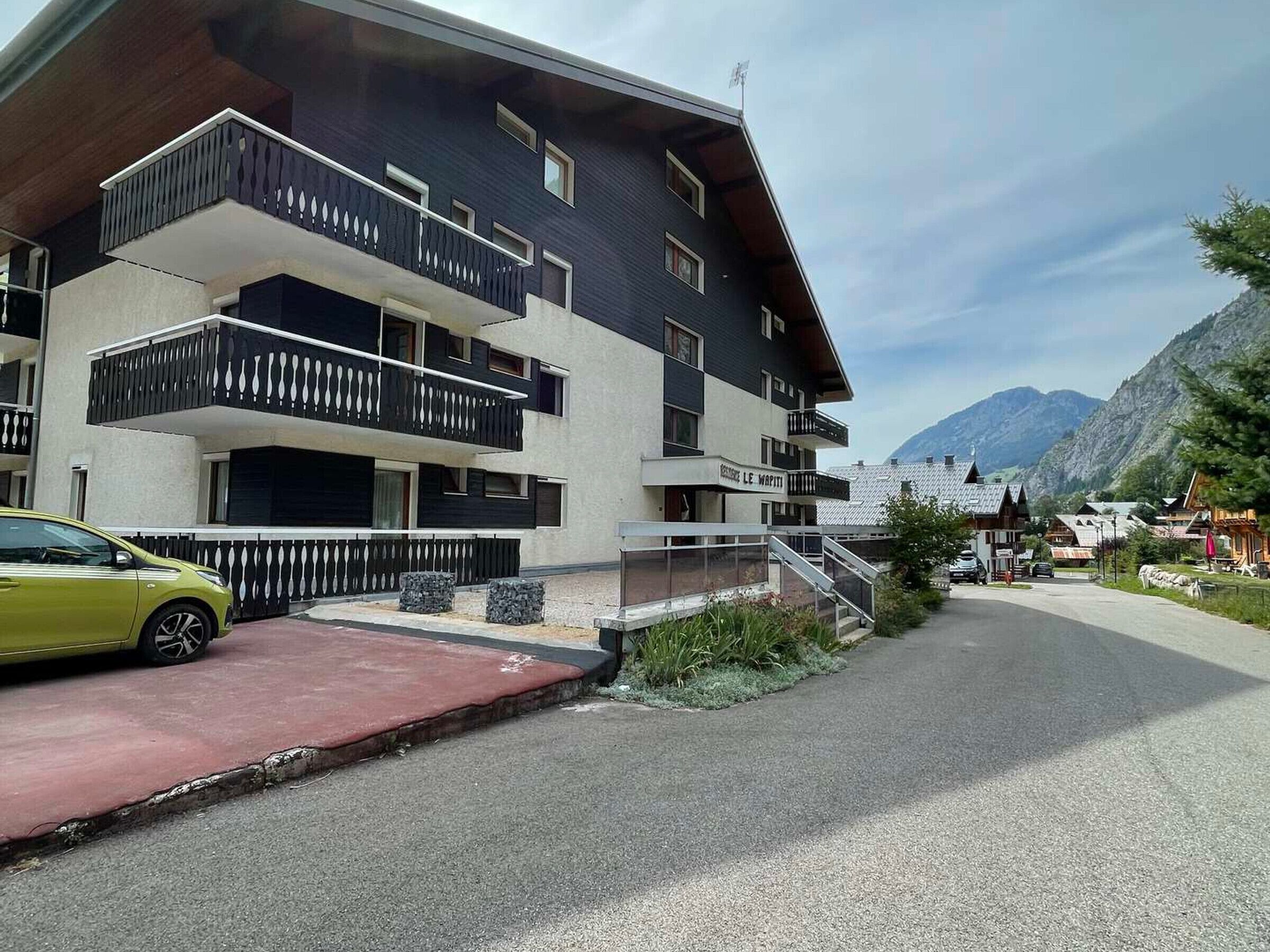 Un edificio moderno de varios pisos con balcones, un coche estacionado al frente y montañas al fondo.