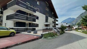 Un immeuble moderne à plusieurs étages, doté de balcons, avec une voiture garée devant et un paysage montagneux en arrière-plan.
