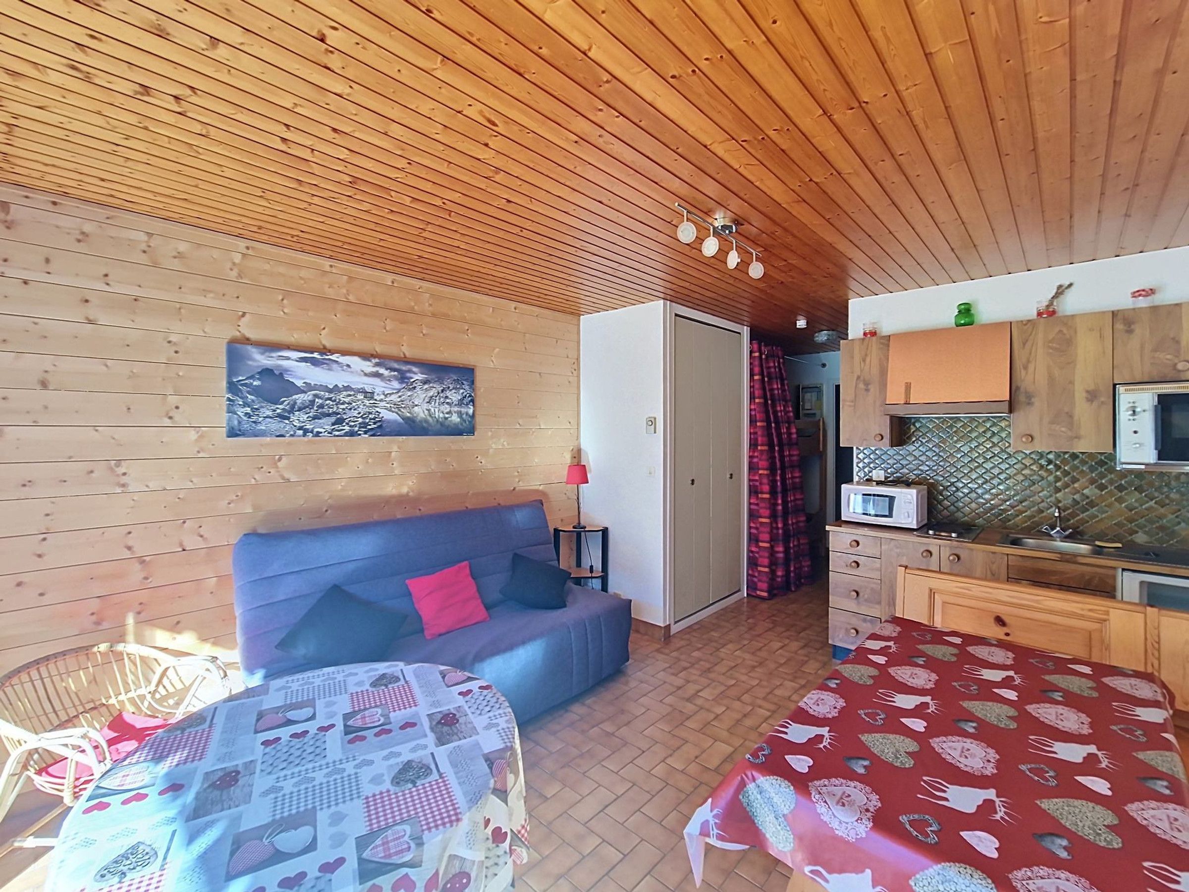 A cozy room with a wooden ceiling, a sofa with a red pillow, a table with a patterned tablecloth, and a kitchenette with a microwave.