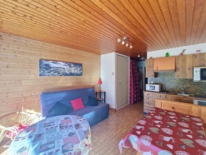 A cozy room with a wooden ceiling, a sofa with a red pillow, a table with a patterned tablecloth, and a kitchenette with a microwave.