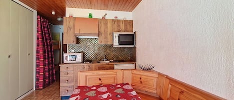 A compact bedroom with a wooden ceiling, a bed with a red patterned bedspread, a kitchenette with a microwave, and a small window.