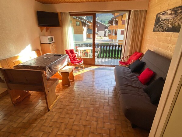 A cozy room with a sofa, a dining table, a TV, and a view of buildings through a glass door. - Cozy studio at the slopes with parking (La Chapelle-d'Abondance)