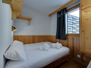 A bedroom with a wooden headboard, a bed with white linens, a window with a view of buildings, and a wooden shelf.