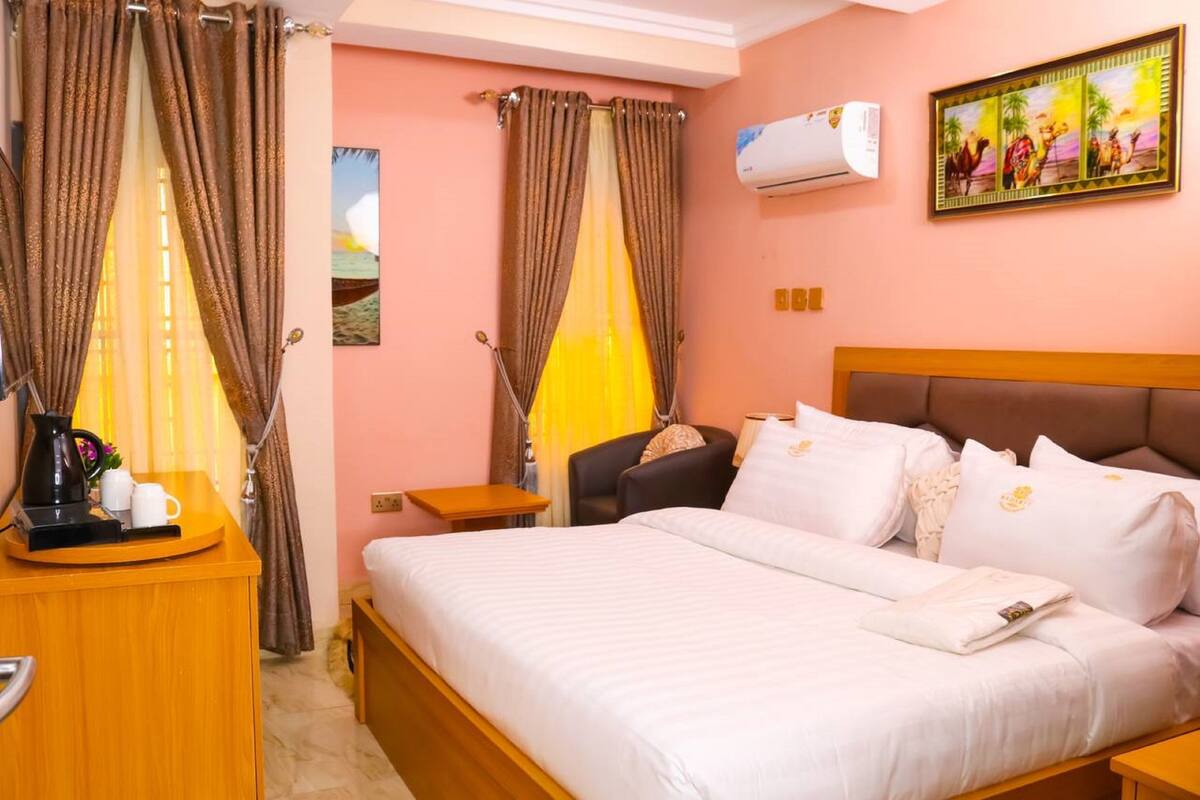 Habitación de hotel con cama, una silla, mesita de noche con cafetera, cortinas, aire acondicionado y un cuadro en la pared.