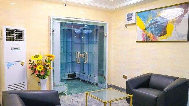 A reception area with a glass door, a white air conditioning unit, a vase of flowers, two black leather chairs, a glass coffee table, and a large abstract painting on the wall. - Madaki's Executive Suites (Abuja)