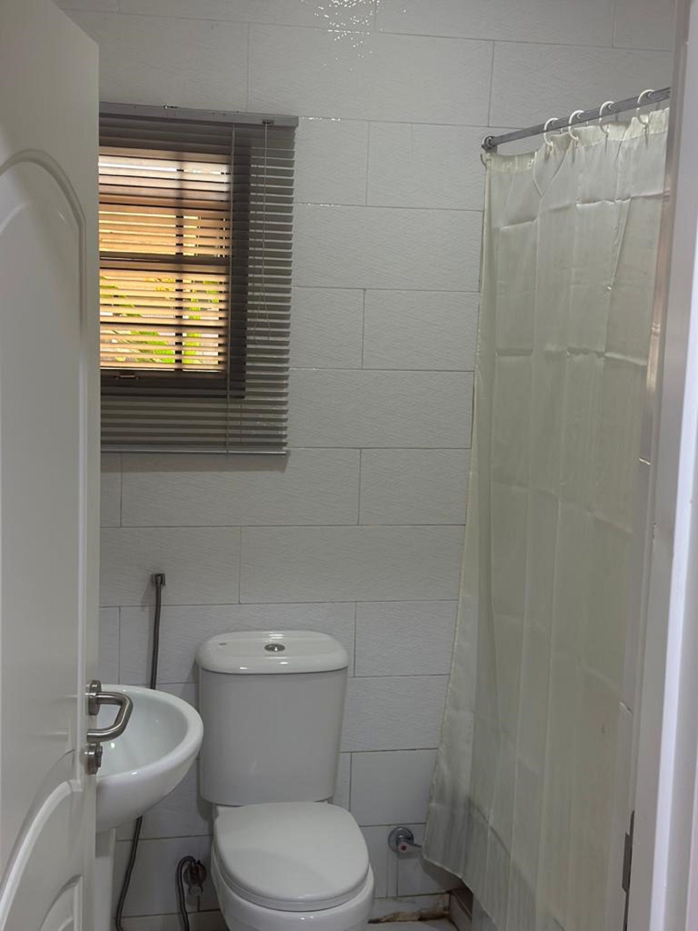 Une petite salle de bain blanche, équipée de toilettes, d’un lavabo et d’une fenêtre avec des stores.