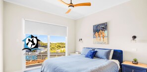 A bedroom with a bed, a ceiling fan, a painting, and a view of the outdoors.