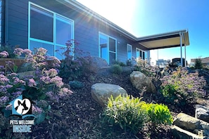 Ein modernes Haus mit großen Fenstern, einer überdachten Terrasse und einem angelegten Garten mit Steinen und Pflanzen.