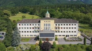 A multi-story building with a central entrance, surrounded by greenery and a parking area. - Kloster by bsmart (Schaan)
