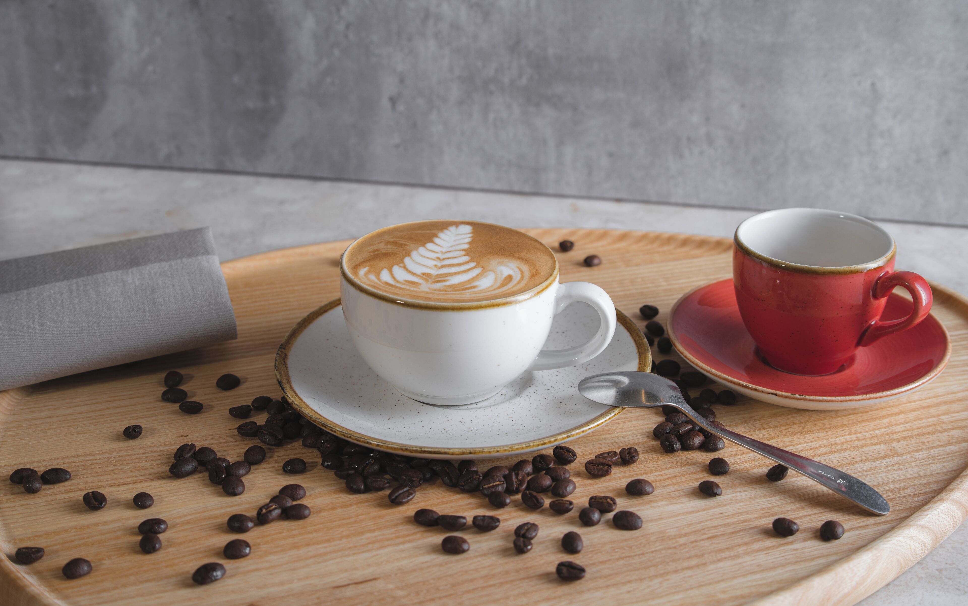 Een witte kop met latte-art op een houten dienblad, een rode kop op een schotel, en losse koffiebonen.