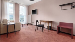 A hotel room with a sofa, a small table, a chair, a desk, a TV, and a window with a view. - Kloster by bsmart (Schaan)