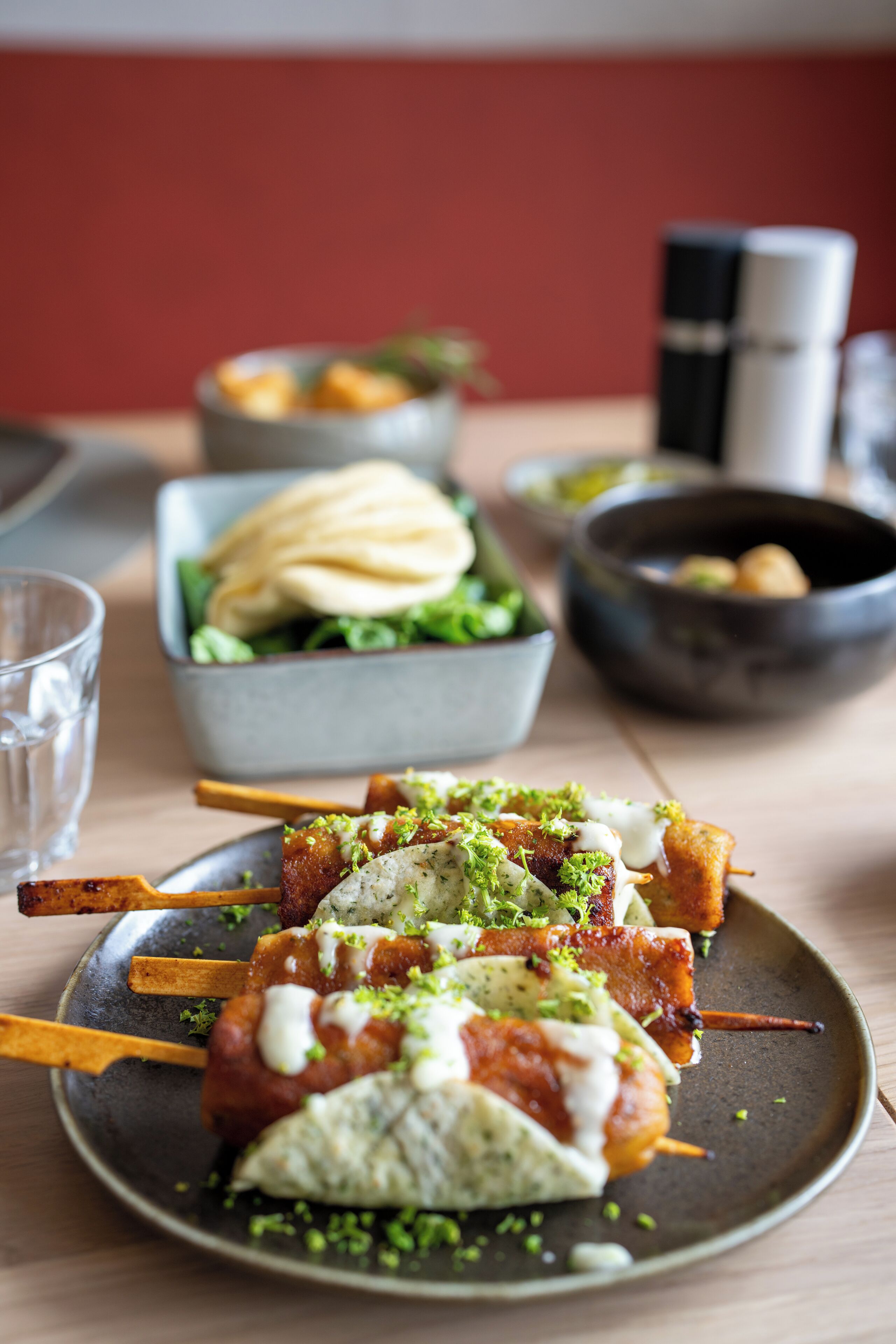 A plate of skewered meat with a creamy sauce and green garnish, accompanied by a bowl of flatbread and a glass of water on a wooden table.