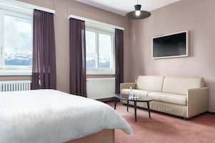 Habitación de hotel con cama, sofá, mesa de centro, televisor y ventana con cortinas.