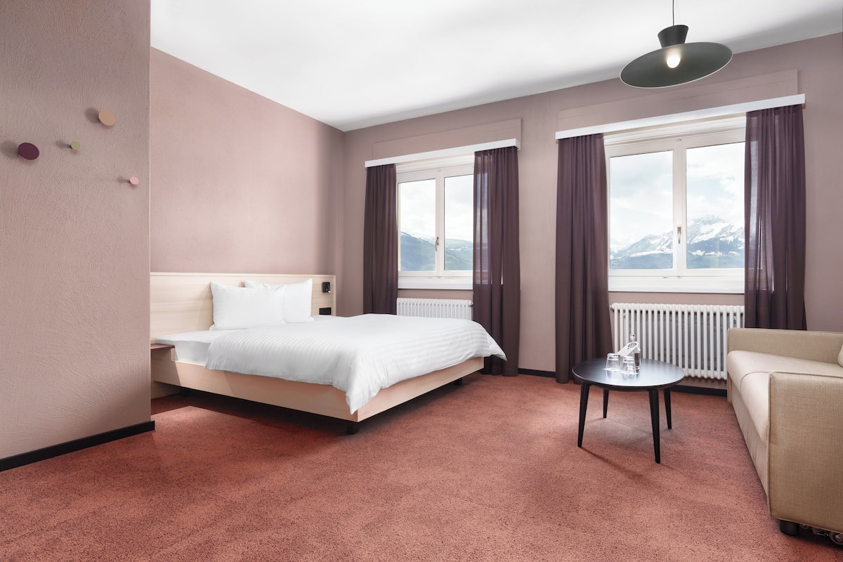 A hotel room with a bed, a sofa, a small table, and a view of mountains through the windows.