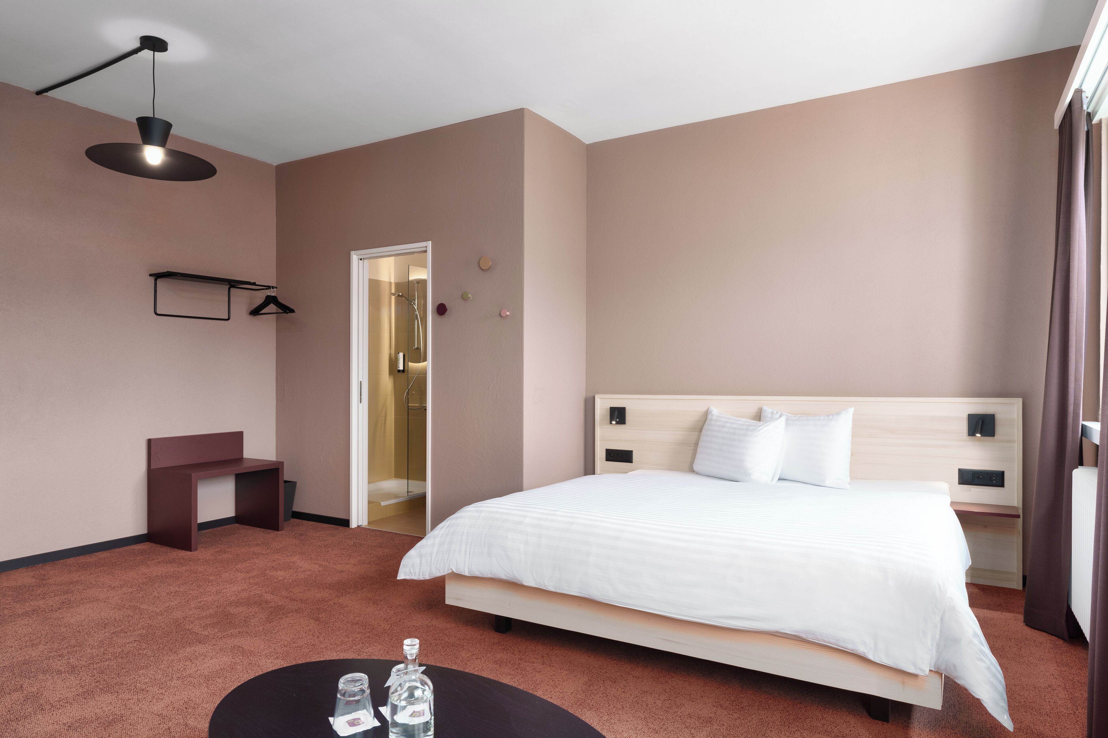 Et moderne hotelværelse med en stor seng, et natbord, et lille skrivebord, en hængende lampe og en dør til et badeværelse.