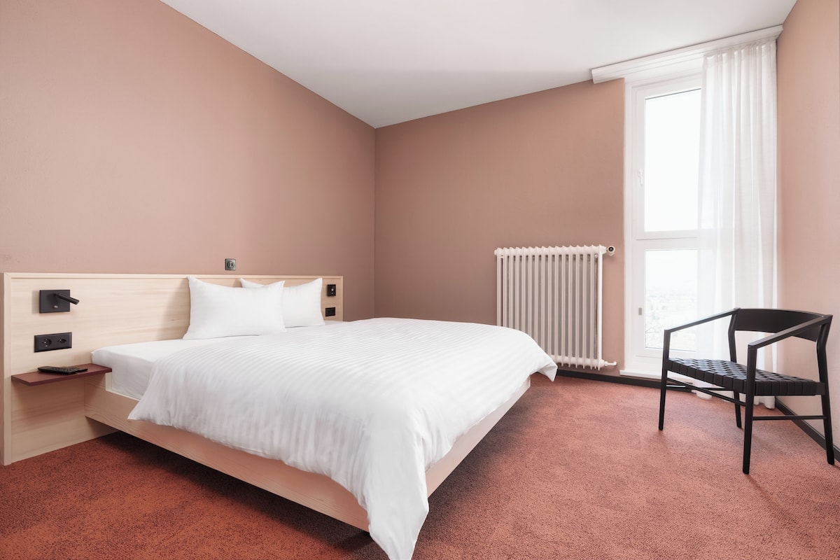 A bedroom with a bed, a chair, a nightstand, a radiator, and a window with curtains.