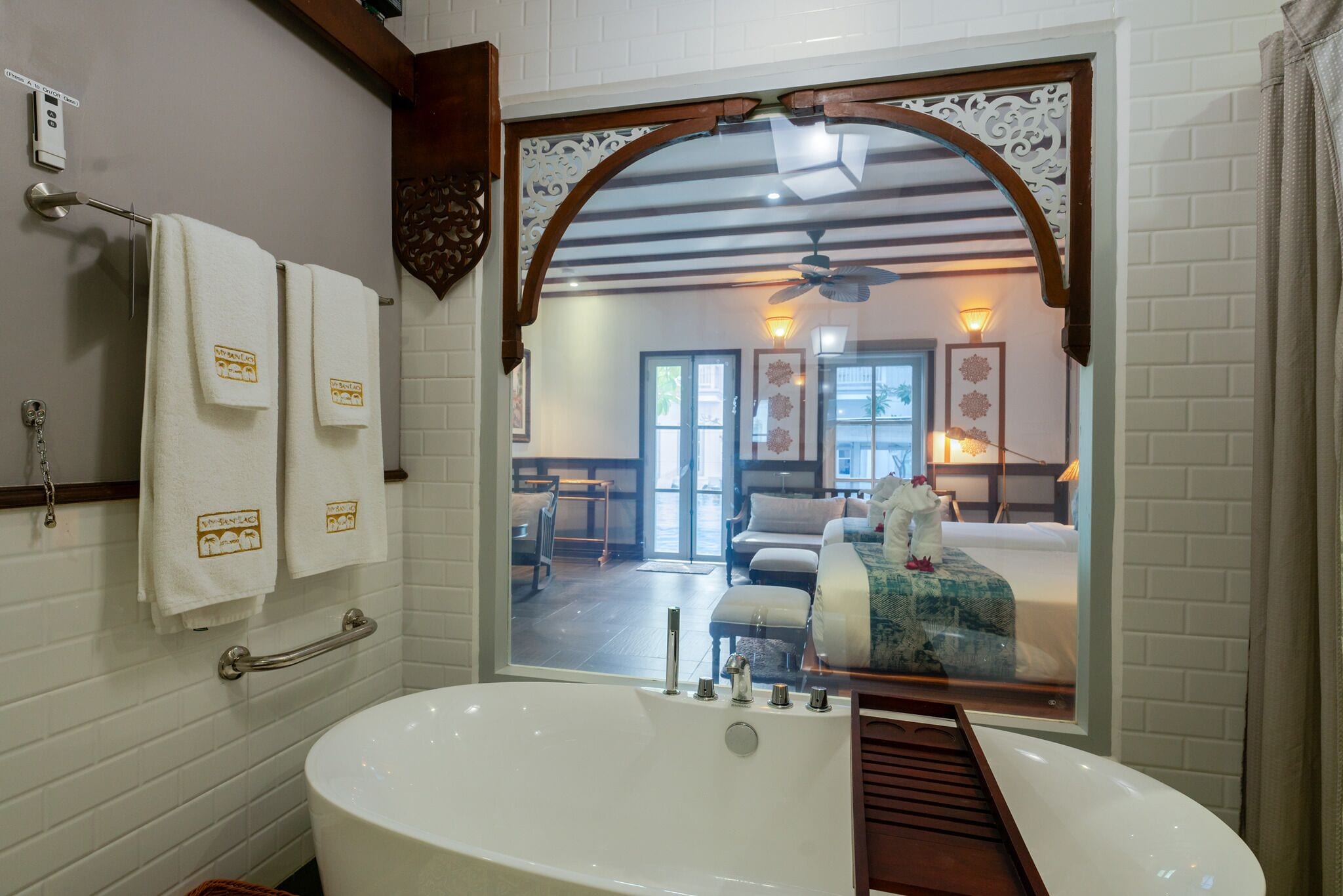 Un baño con una bañera grande, un perchero con dos toallas, un pasamanos y un espejo que refleja un dormitorio con una cama y una ventana.