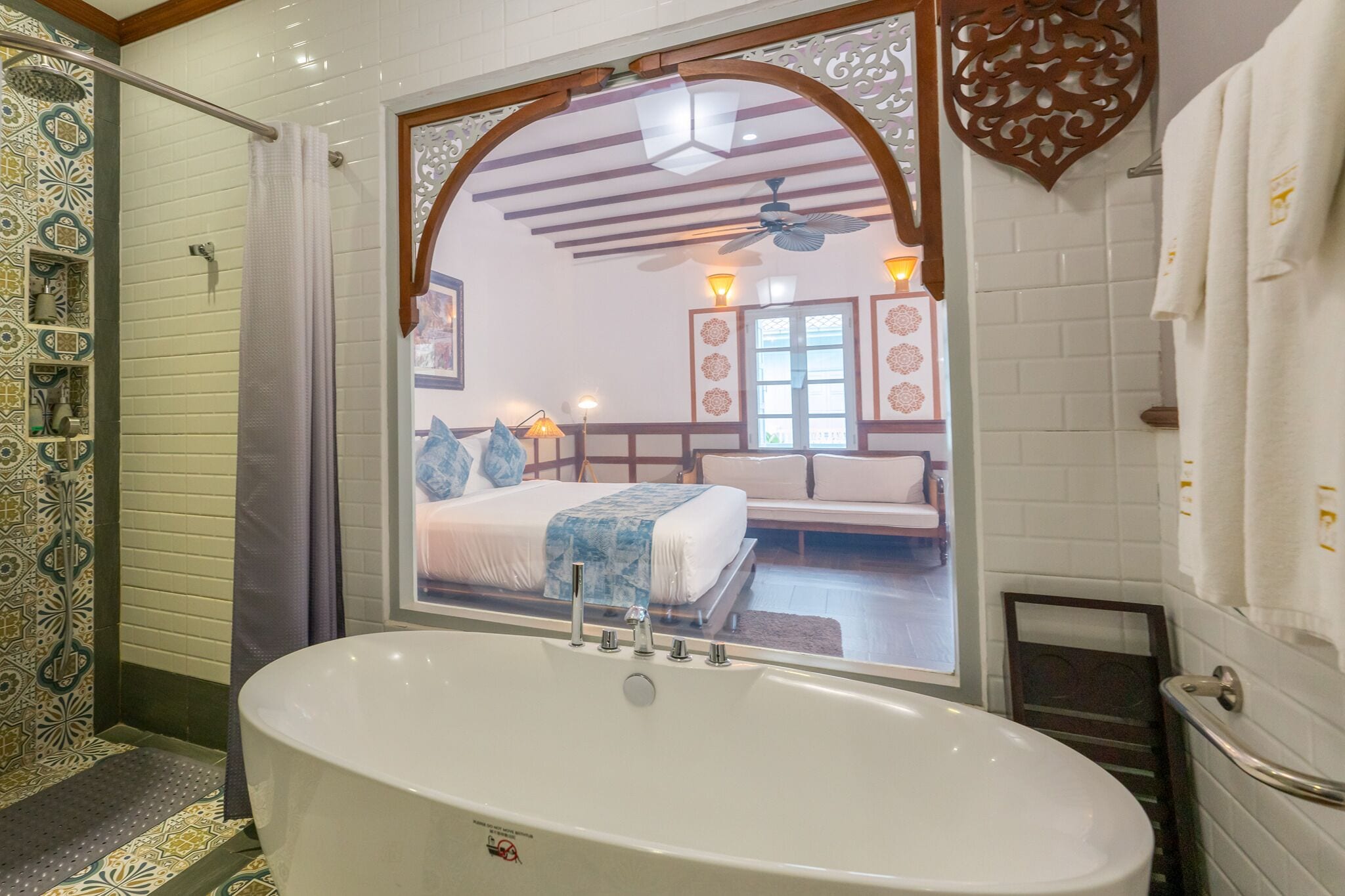 Un baño con una bañera grande, zona de ducha y un espejo que refleja un dormitorio con cama y ventana.