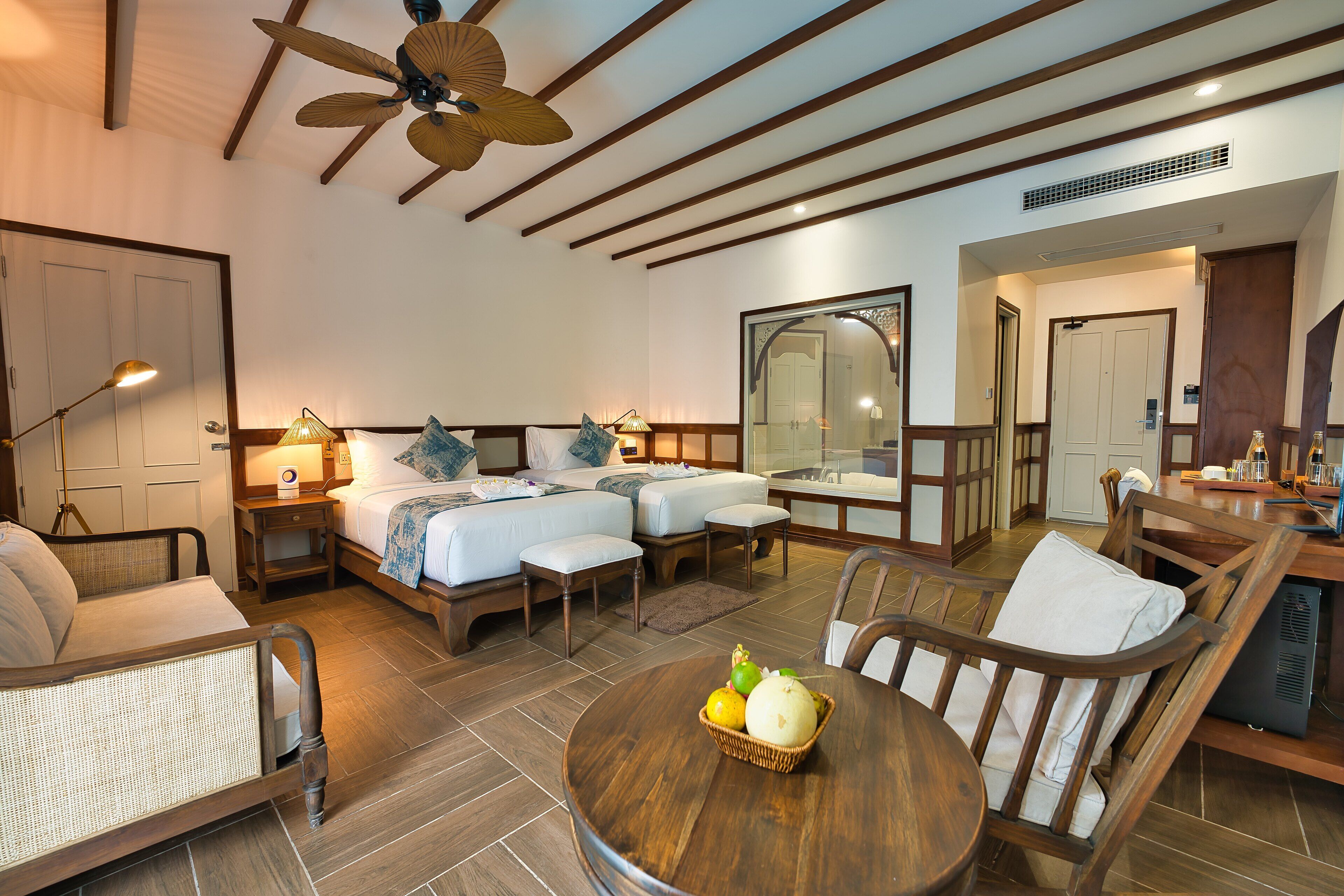 A hotel room with a bed, two bedside tables, a chair, a dining table with a fruit basket, and a ceiling fan.