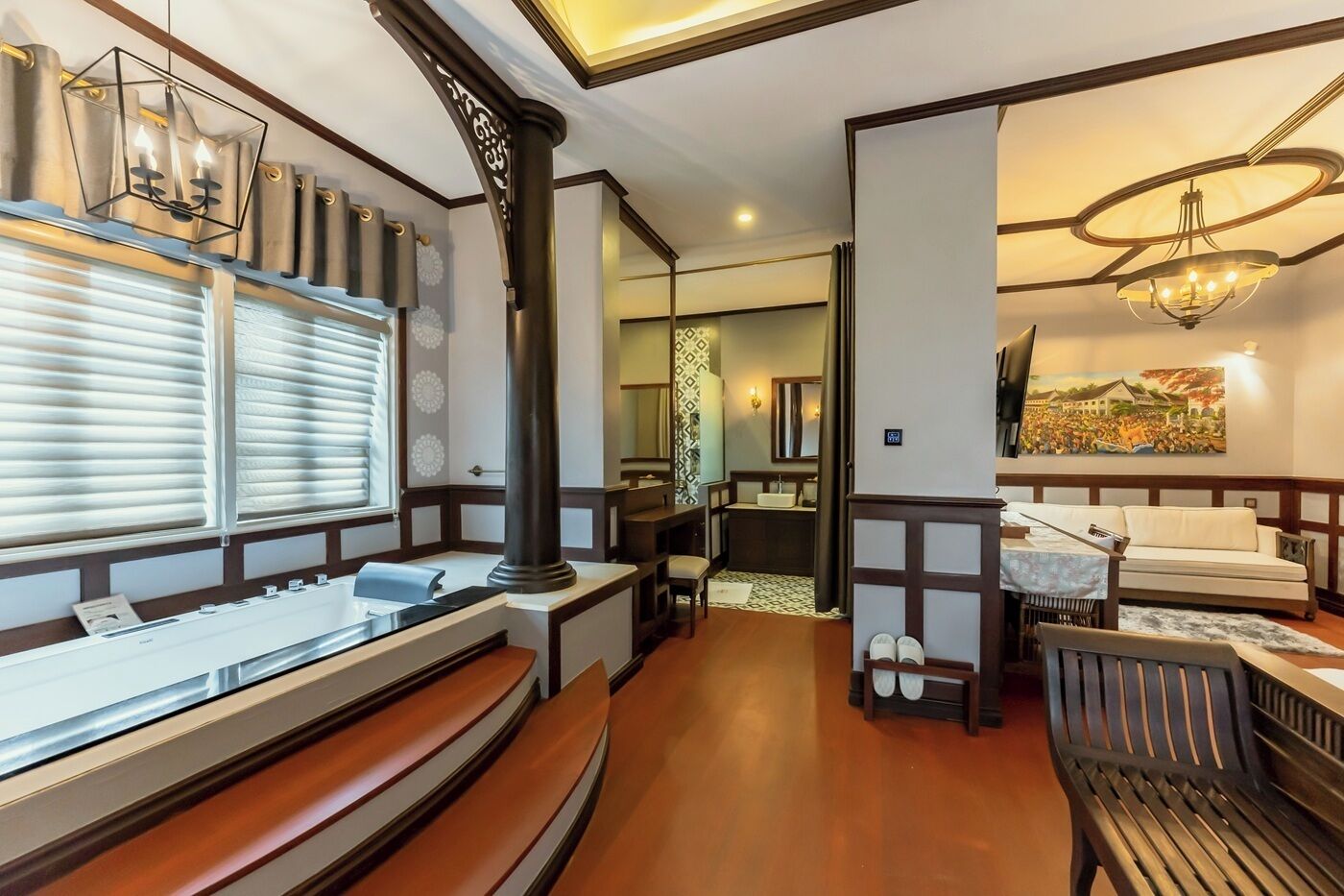 A hotel room with a wooden floor, a large window with blinds, a desk with a chair, a dining area with a table and chairs, and a bathroom with a sink and mirror.