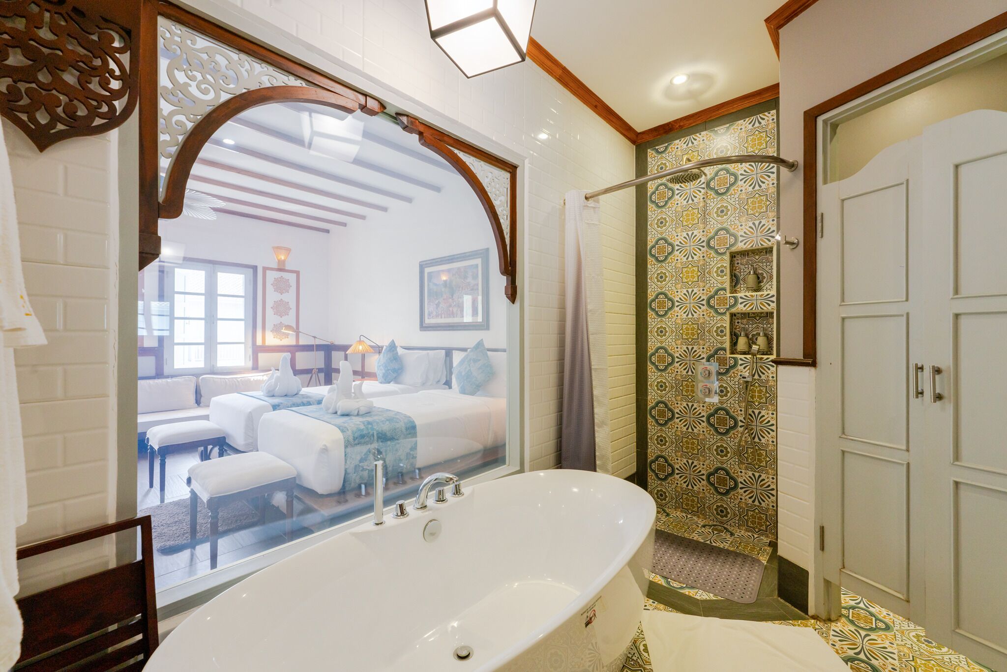 A modern bathroom with a freestanding bathtub, a shower area with patterned tiles, and a view into a bedroom with a large bed and a nightstand.