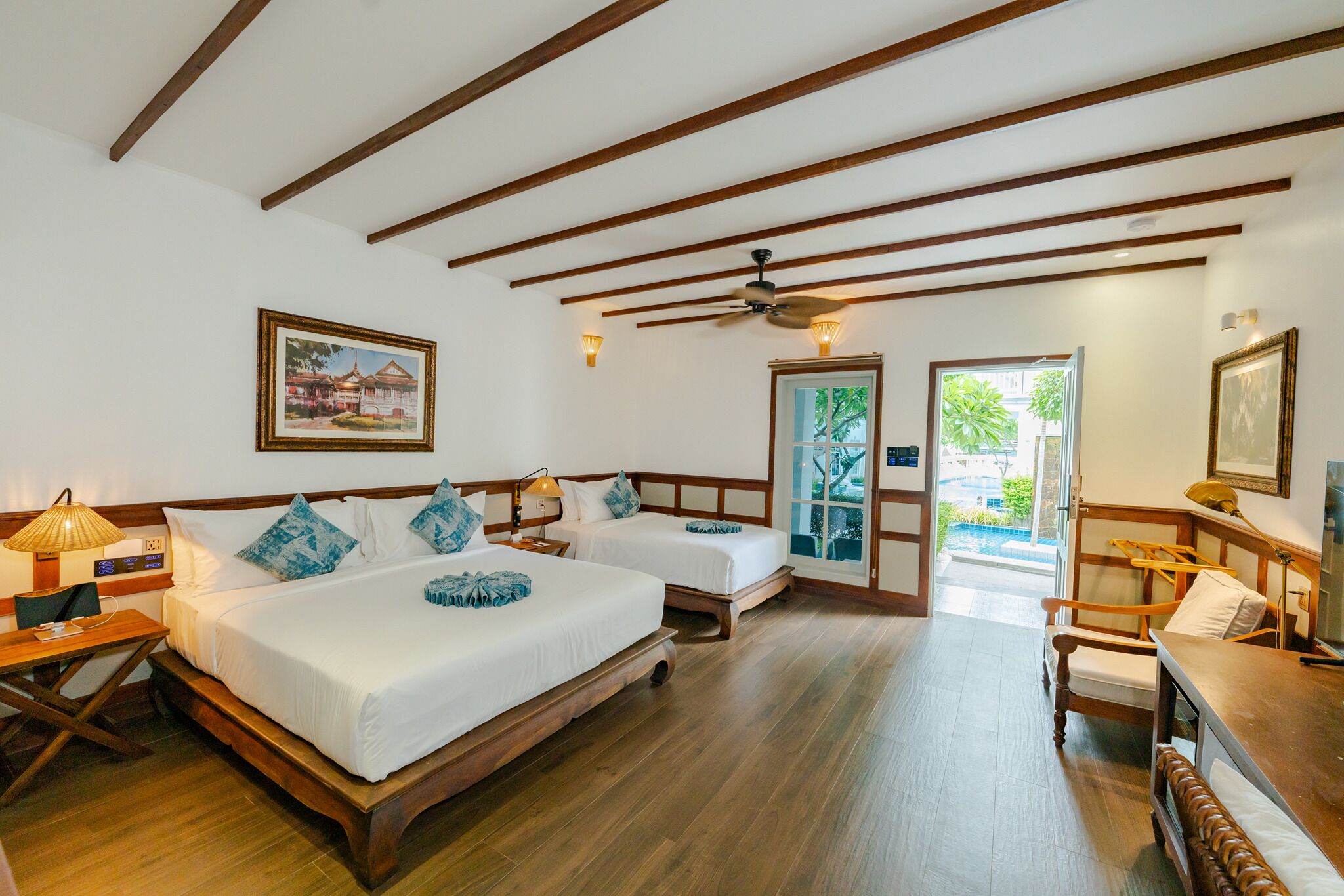 A hotel room with two beds, wooden floors, a ceiling fan, and a view of greenery outside.