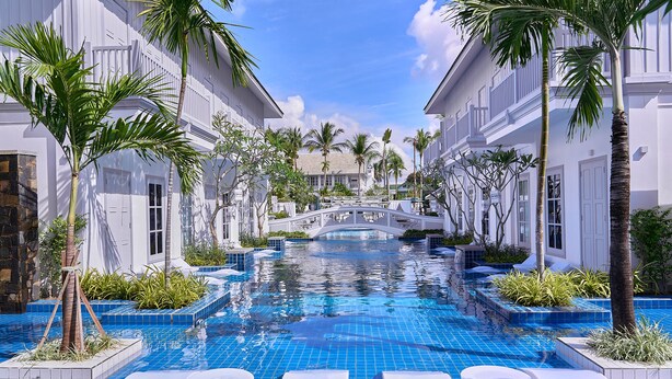 Un área de piscina con un puente, palmeras y edificios blancos.