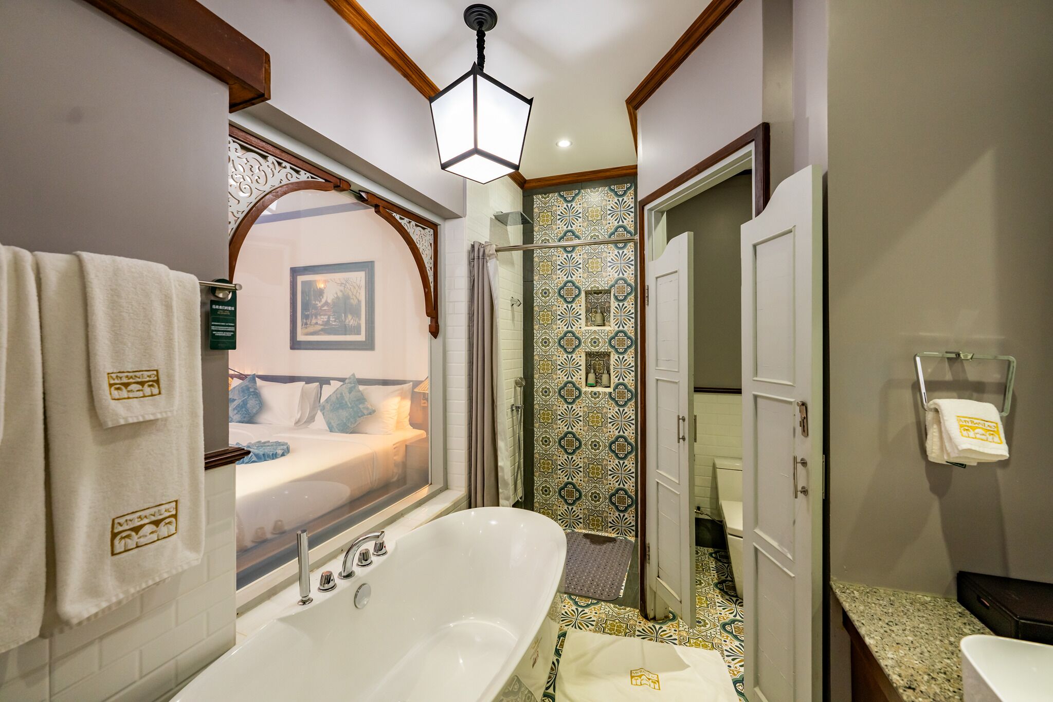 Un baño moderno con una bañera independiente, ducha con baldosas estampadas y una ventana por la cual se ve una cama.