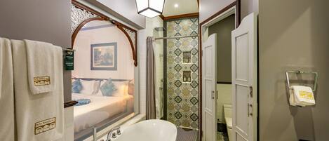 Un baño moderno con una bañera independiente, ducha con baldosas estampadas y una ventana por la cual se ve una cama.