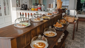 Montaje de bufé con distintos alimentos cubiertos por cúpulas de vidrio, una estación de café y un área de asientos con piso estampado.